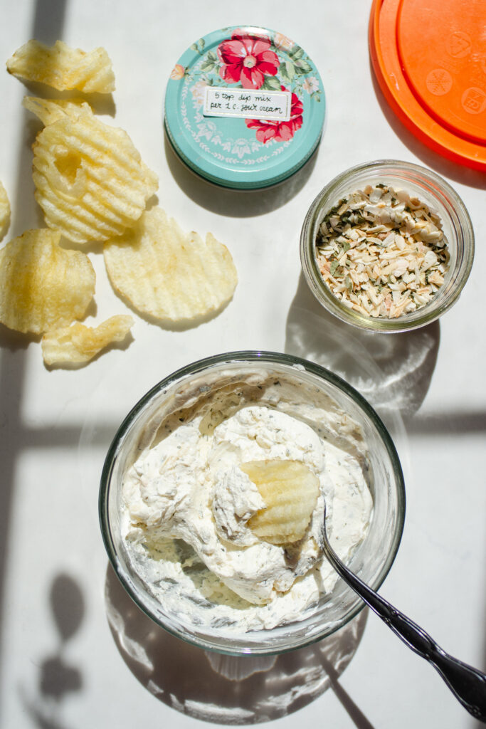 The finished onion dip in a glass storage container with the lid, jar of the onion dip mix, and a few potato chips on the counter beside it.