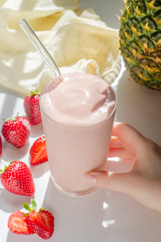 A strawberry pineapple smoothie on a sunny countertop with fresh strawberries and a pineapple in the background. 
