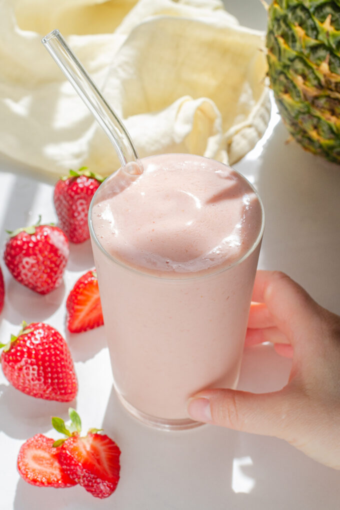 A strawberry pineapple smoothie in a glass with a glass straw with fresh strawberries on one side, a whole pineapple in the background, and a hand holding the glass.