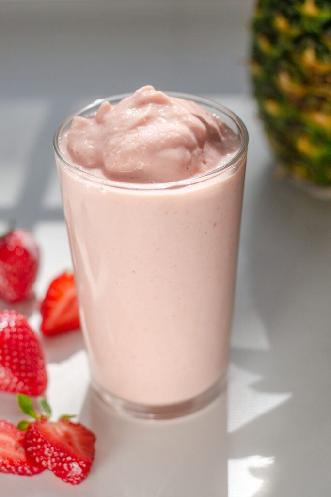 The finished strawberry pineapple smoothie in a glass with fresh strawberries off to the side and a whole pineapple on the other side.