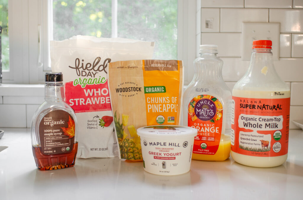 All of the ingredients needed to make a strawberry pineapple smoothie laid out on a white countertop with a window in the background. 