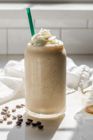 A Starbucks copycat Coffee Frappuccino in a glass cup on a white countertop with espresso beans on one side and a white napkin draped in the background.
