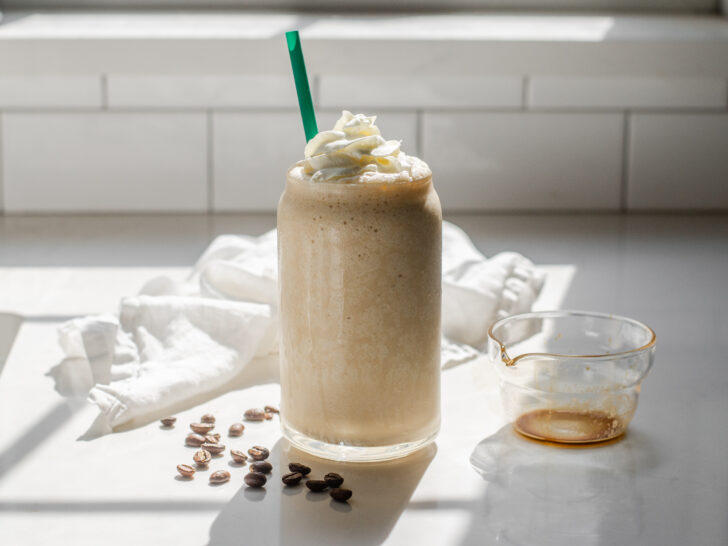 A Starbucks copycat Coffee Frappuccino in a glass cup on a white countertop with espresso beans on one side and a white napkin draped in the background.