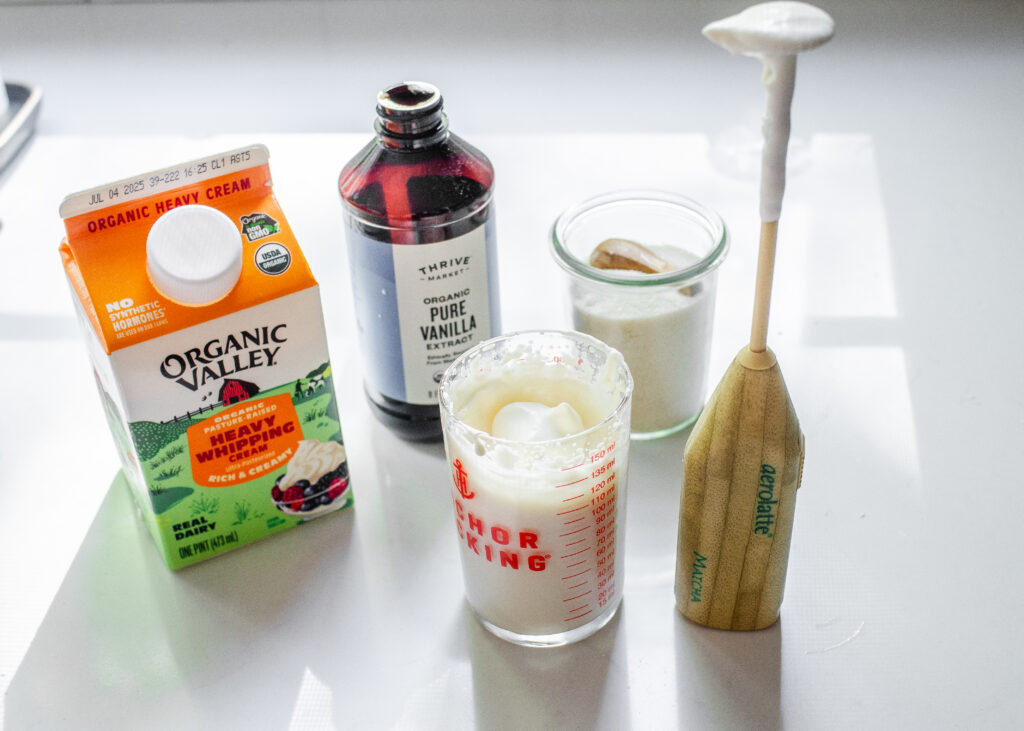 A photo showing all of the ingredients needed to make a small batch of homemade whipped cream, with the finished whipped cream in a small glass.
