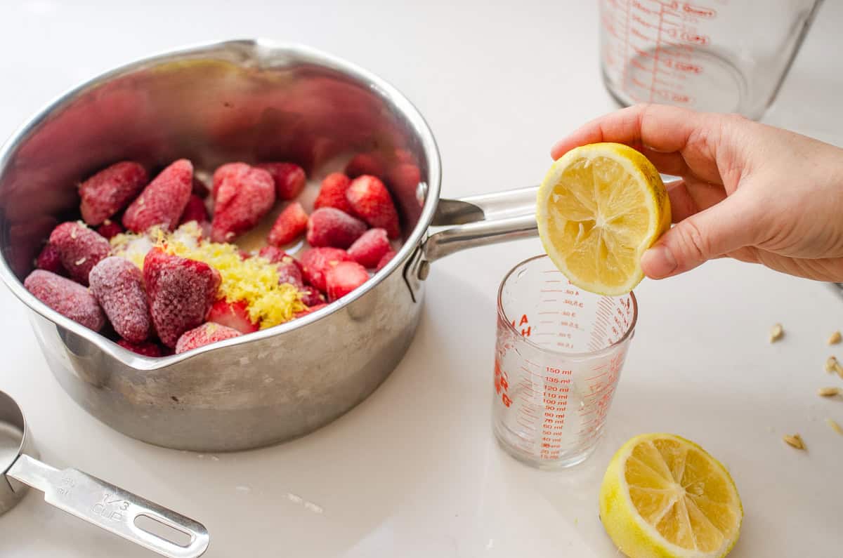 Squeezing a lemon into a small measuring cup with the pan of strawberries off to the side.