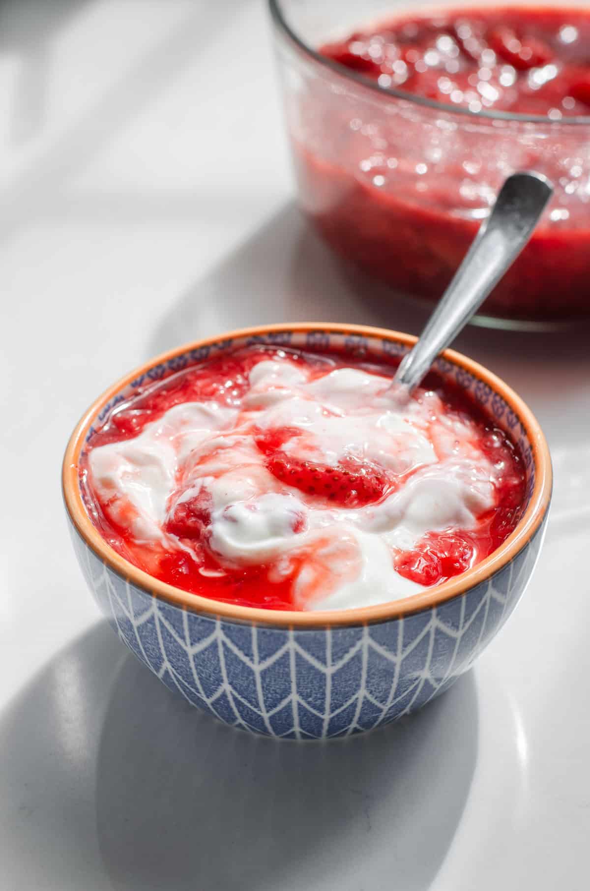 A small blue bowl of Greek yogurt with a spoon in it with strawberry sauce partially mixed in, and the bowl of sauce in the background.
