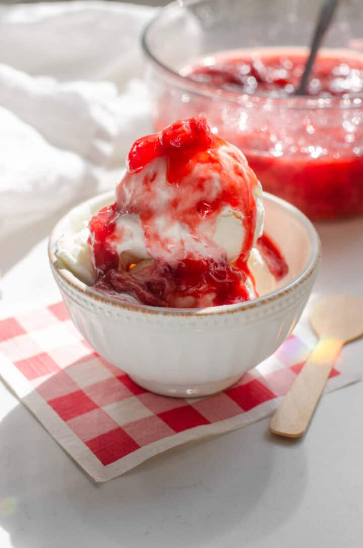 A bowl of vanilla ice cream with strawberry sauce on top with the bowl on a red and white gingham napkin with a wooden spoon off to the side.