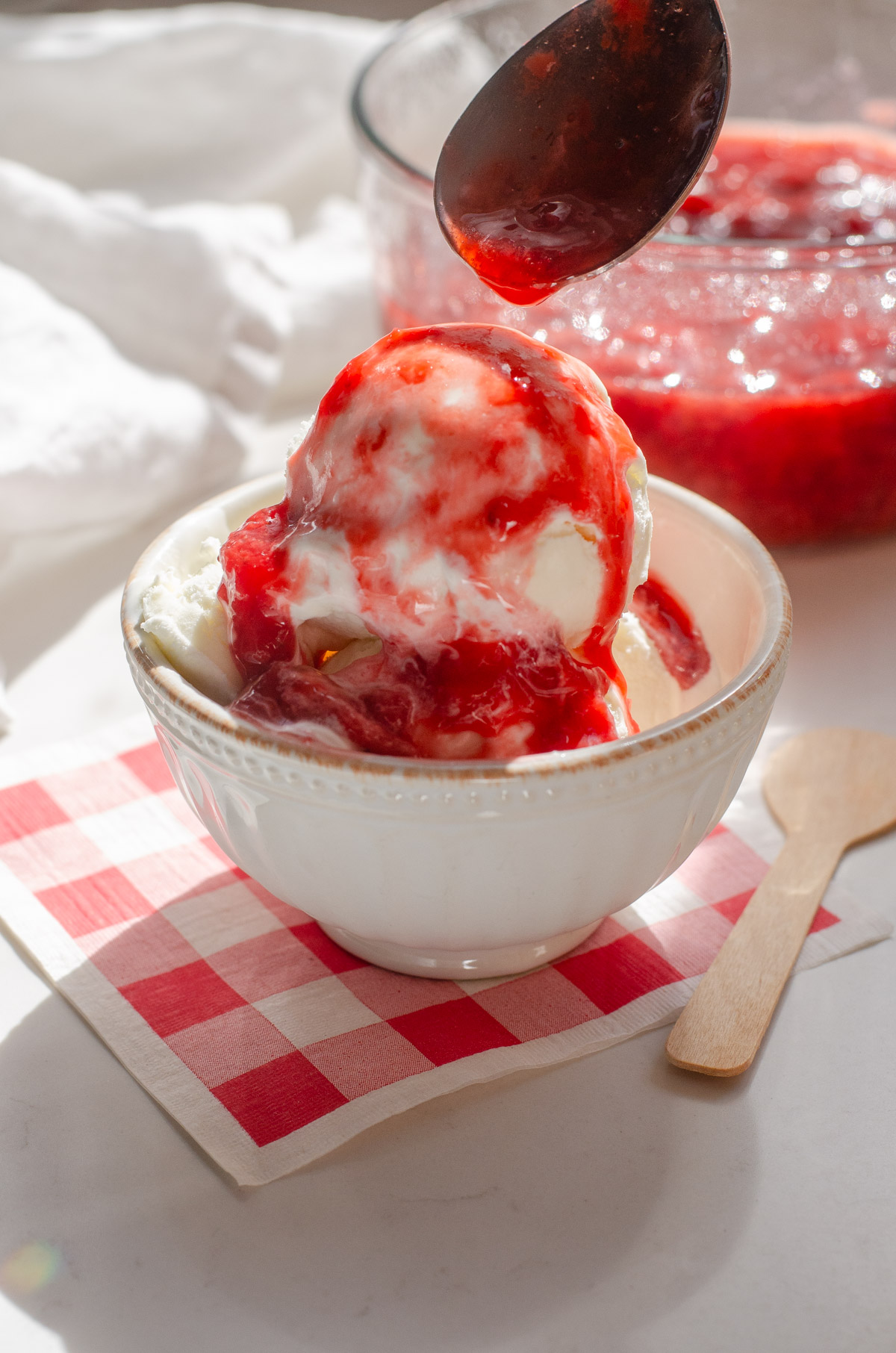 A bowl of ice cream on a gingham paper napkin with a spoon adding strawberry sauce on top.