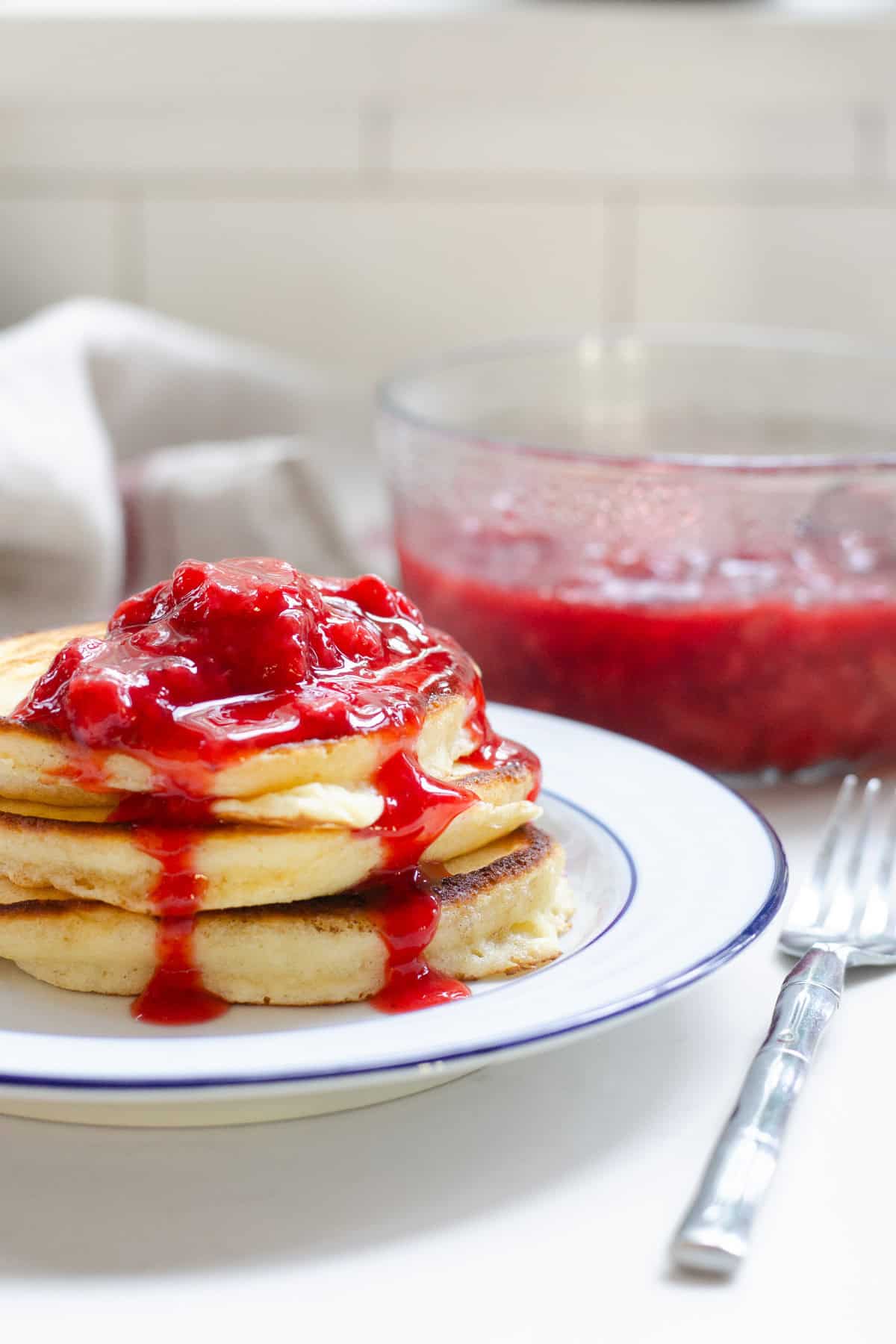 A stack of pancakes on a white plate with a blue rim with strawberry sauce on top of the pancakes and dripping down the sides. The bowl of sauce is in the background, as well as a linen tea towel.