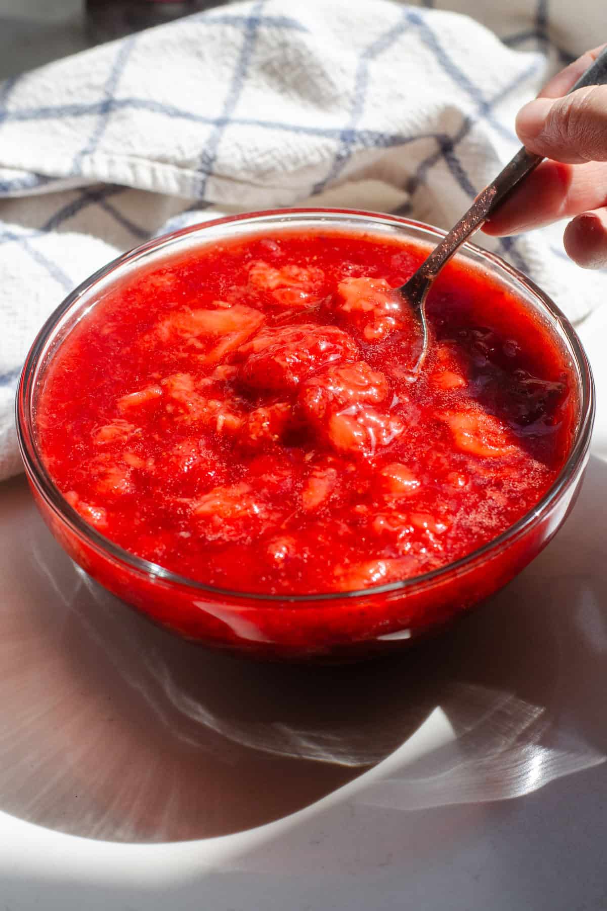 A bowl of strawberry sauce on a sunny countertop with a hand lifting a spoon and a checkered tea towel in the background.