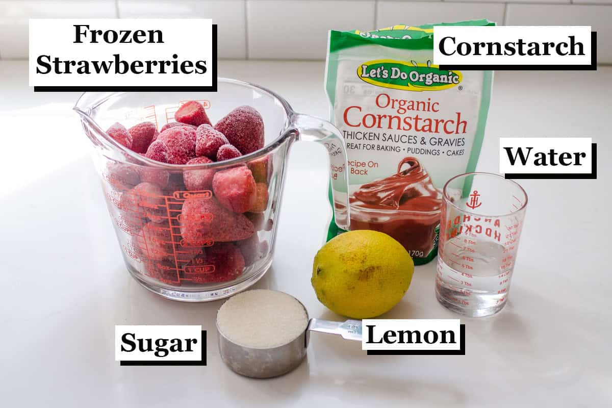 Frozen strawberries, cornstarch, water, a whole lemon, and sugar laid out on a white countertop with text labeling the ingredients.