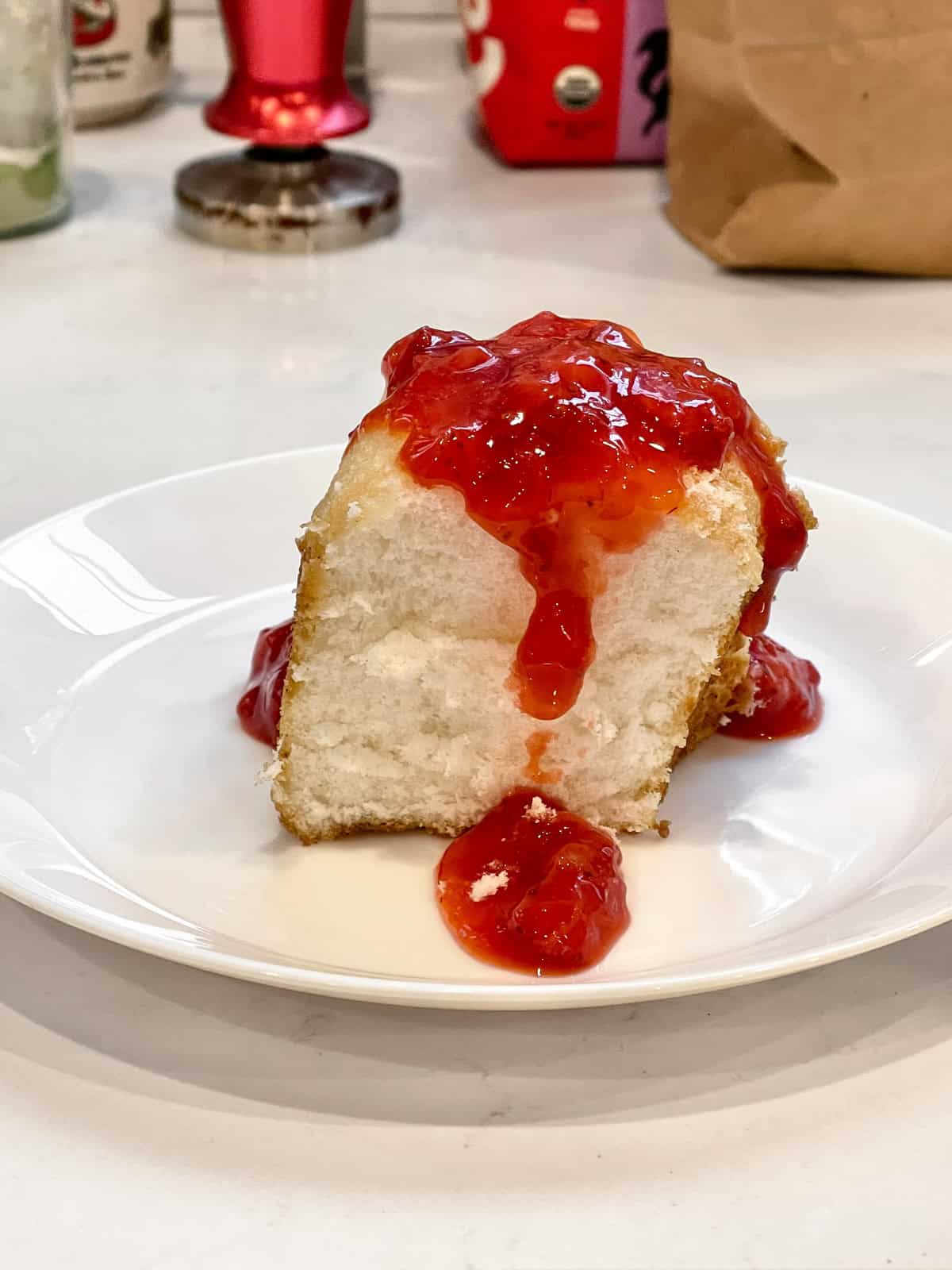 A slice of Angel Food Cake on a white plate with strawberry sauce on top and dripping down the side.