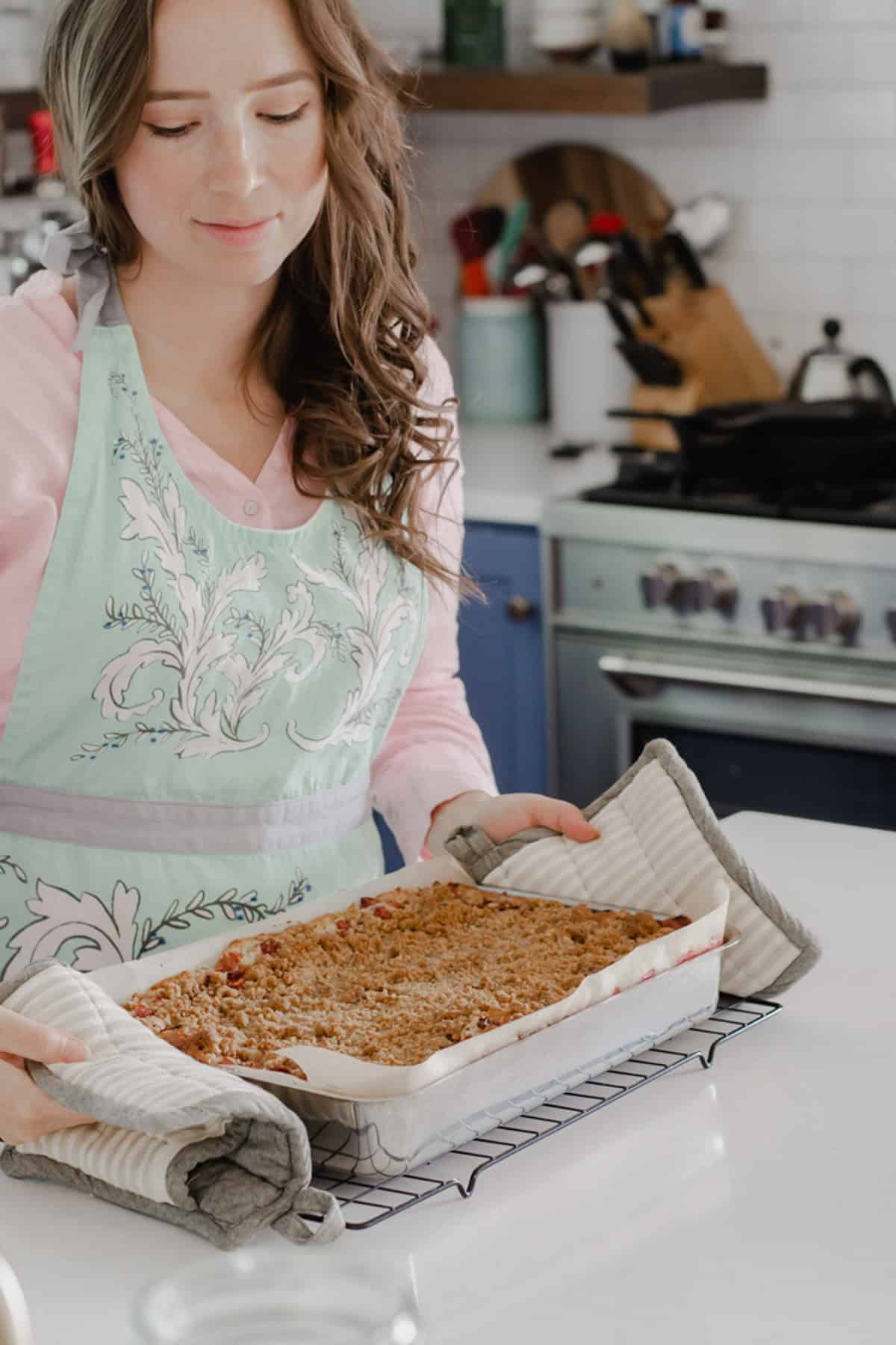 A woman in an apron holding a 13x9 inch pan with hot pads.