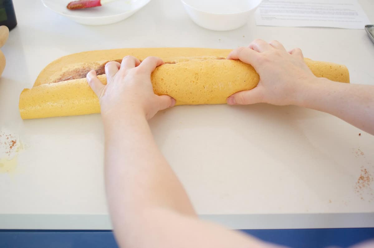Two hands rolling up the pumpkin cinnamon roll dough jelly-roll style.