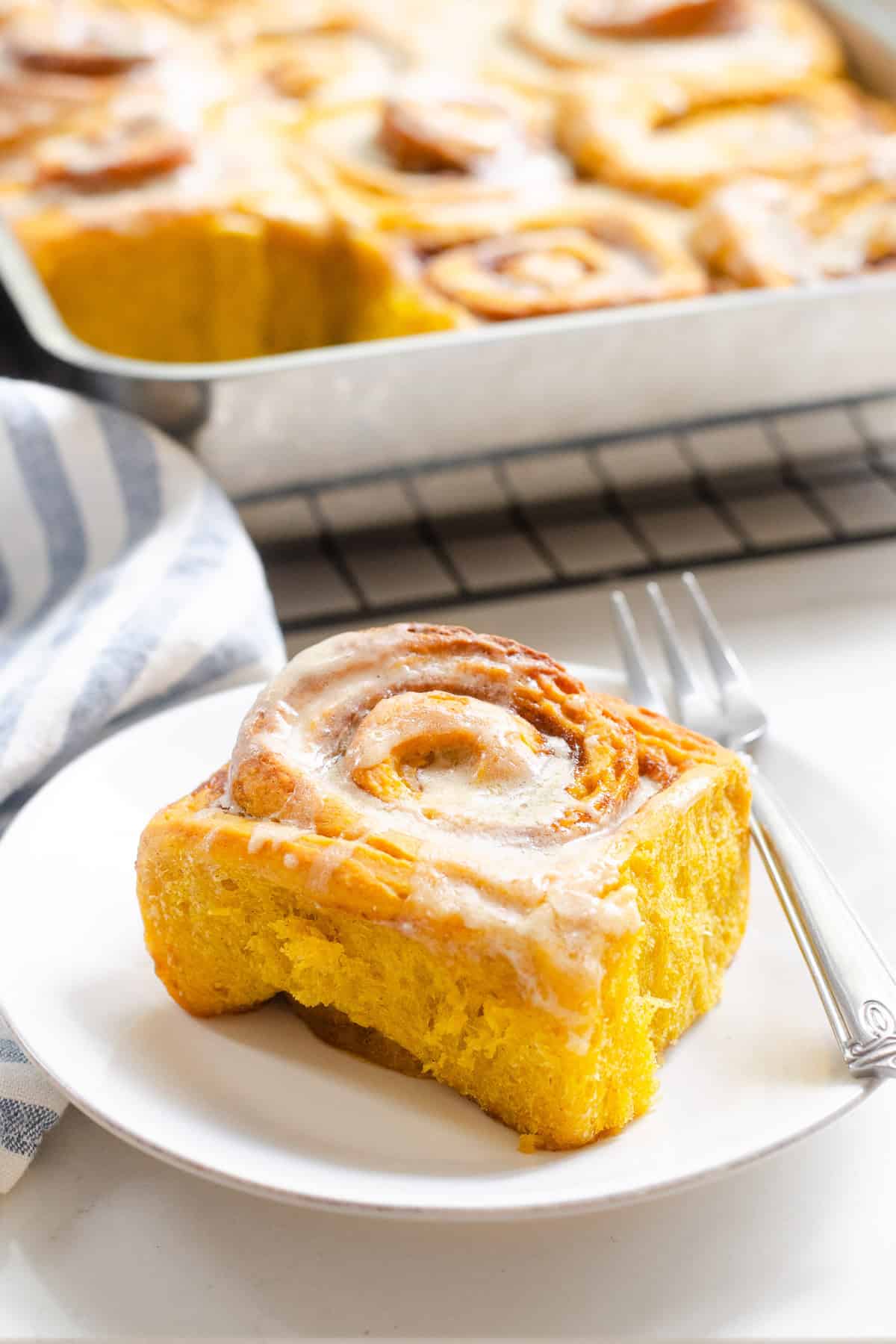 A single sourdough pumpkin cinnamon roll on a small plate with a dessert fork to the side, and the pan on rolls in the background.