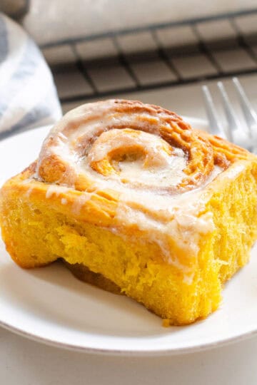 A sourdough pumpkin cinnamon roll on a white plate with a dessert for on the right and a striped linen tea towel on the left.